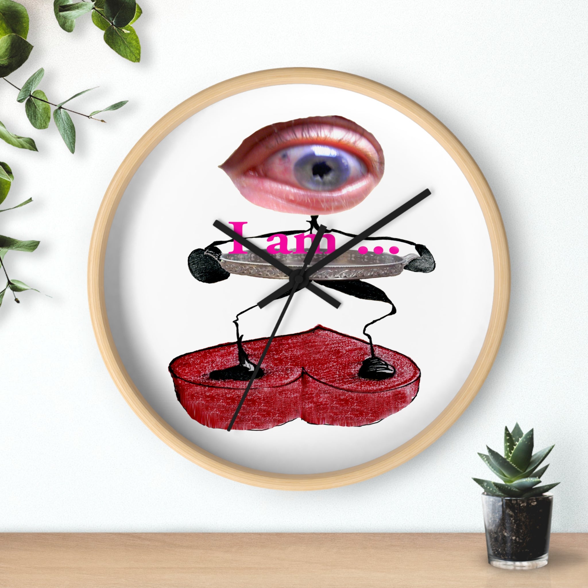 Surreal eye art wall clock with pink eye above “I am...” text, black stick figures holding a red heart platform on a white face in a light wood frame, on a shelf beside a small plant.