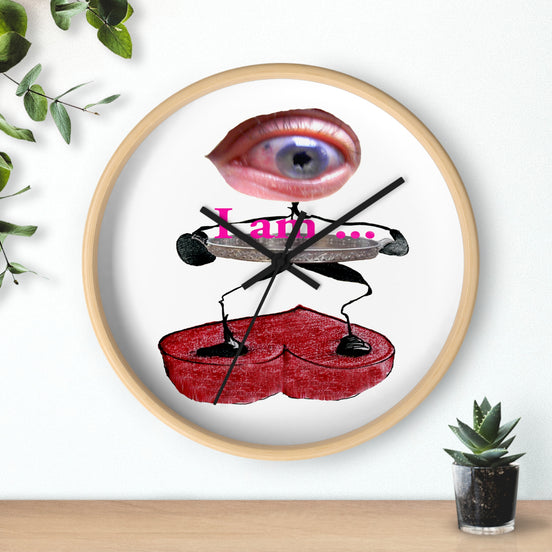 Surreal eye art wall clock with pink eye above “I am...” text, black stick figures holding a red heart platform on a white face in a light wood frame, on a shelf beside a small plant.